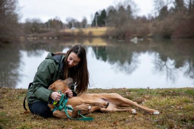 Woman in green jacket with tan colored dog by a pond