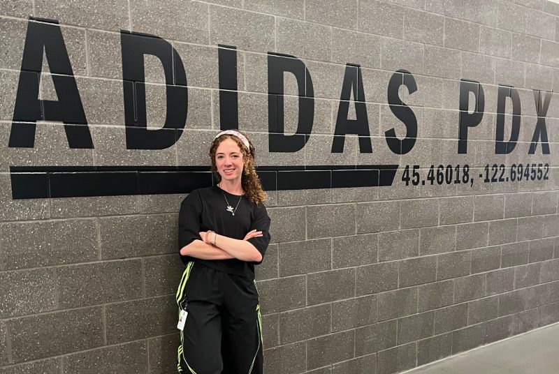 A student stands smiling with arms crossed in front of a gray brick wall displaying large black letters that read “ADIDAS PDX” and geographic coordinates.