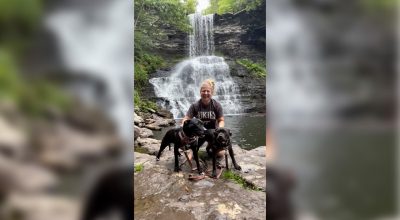 Person posing with two black dogs in front of a waterfall.