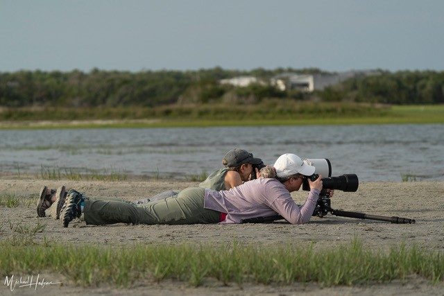 Two people laying down beside water with photography equipment to capture wildlife.