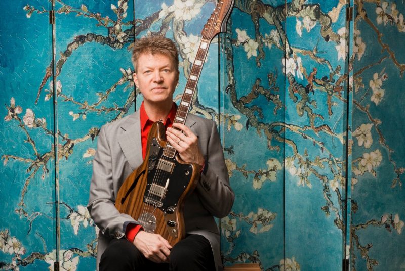 Nels Cline, dressed in a suit with a red shirt, sits holding a guitar in his lap in front of an Asian-inspired backdrop with blooming tree limbs.