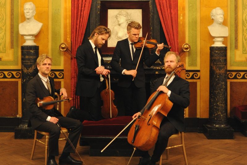 Musicians for the Danish String Quartet pose in an art gallery holding their instruments. Each member is wearing a suit and tie, two are sitting and two are standing in front of a painting and two sculptures.