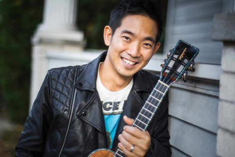 Musician Jake Shimabukuro poses in front of a house in a black leather jacket holding a ukulele.