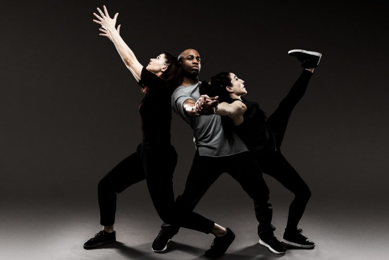 Three dancers all dressed in dark colors pose closely together - one reaching for the sky while the one in the middle holds up the other - in front of a gray background.