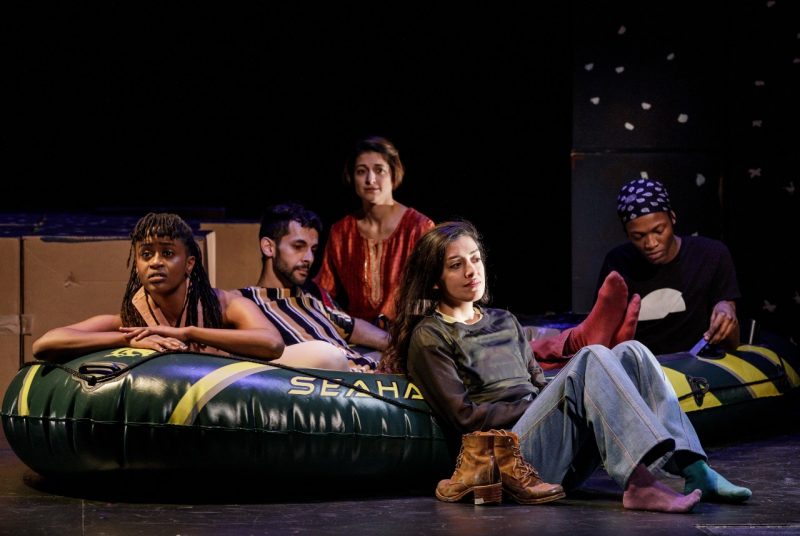 Five young people, members of the cast of the multimedia play "Cartography," sit in an inflatable raft onstage in front of a group of cardboard boxes.