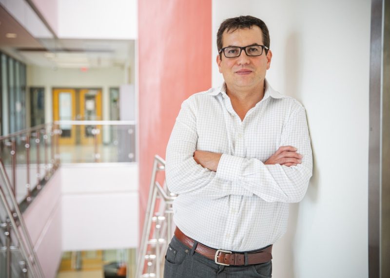 Portrait of Rafael Davalos, standing in Kelly Hall, in a white button-up shirt, black framed glasses, and a slight smile. Standing with crossed arms.