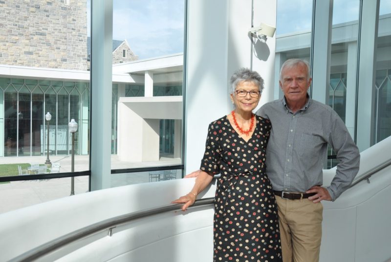 Martha Olson and her husband Thomas pose on a staircase in the Moss Arts Center - the front entrance of the center is visible through large glass windows behind them.