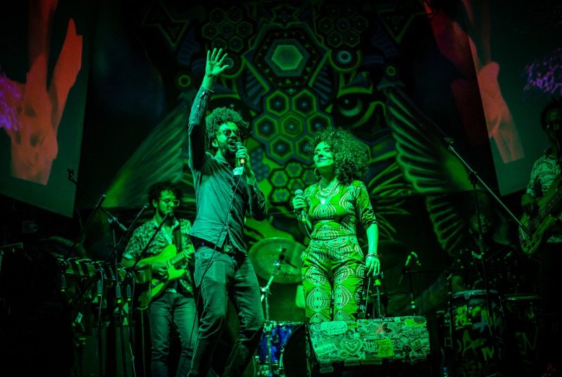 Four members of the band Chontadelia are seen performing onstage, bathed in green light with a backdrop featuring a variety of shapes and designs.