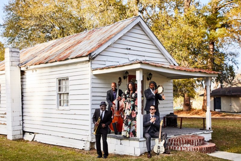 Five musicians with the band Ranky Tanky are on the porch of an old, white house holding their instruments. 
