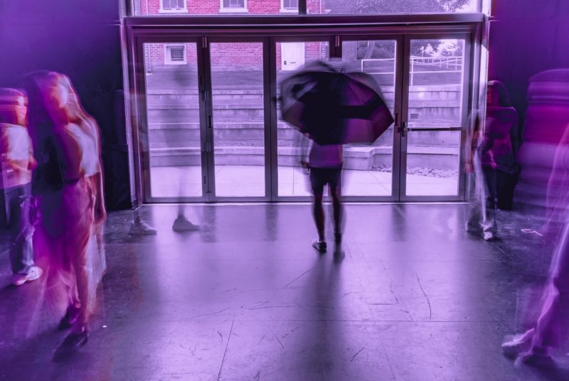 Blurred, purple-pink images of several people walking around a room, one with an umbrella,.