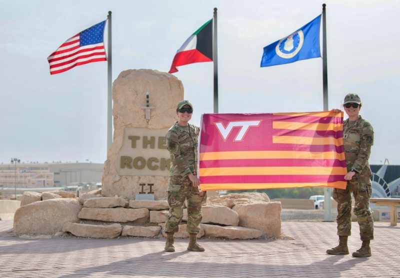 First Lieutenants Abby Houghtling and Colleen Pramenko smile and hold a striped Virginia Tech flag near a monument called “The Rock” in a desert setting.