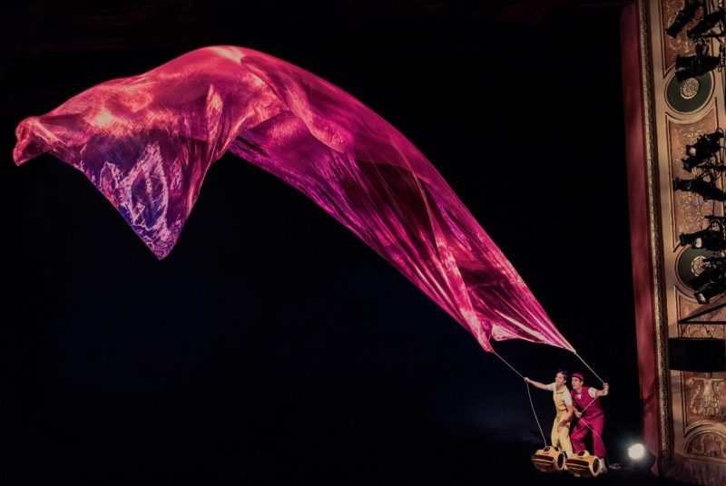 Two performers stand on a platform, each holding strings connected to a rich pink piece of fabric soaring in the air.