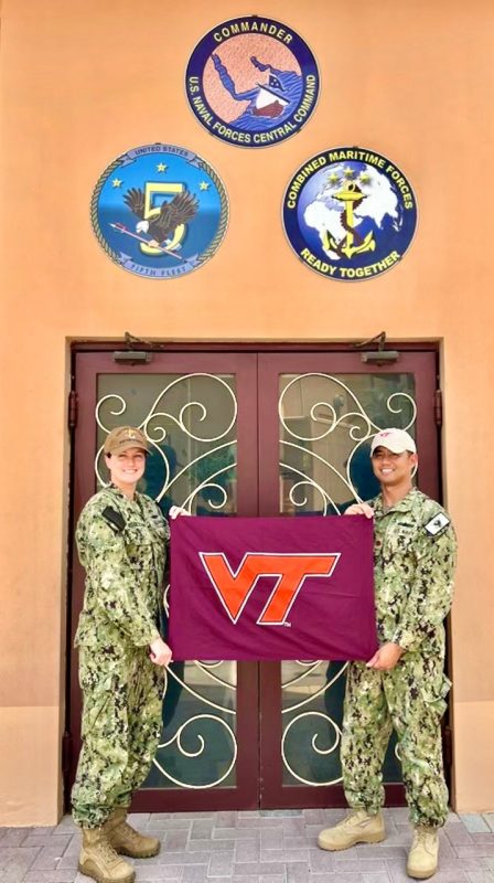 Lieutenant Commander Eggleston and Lieutenant Commander Ignacio stand in camouflage uniforms holding a Virginia Tech flag. Both are smiling.