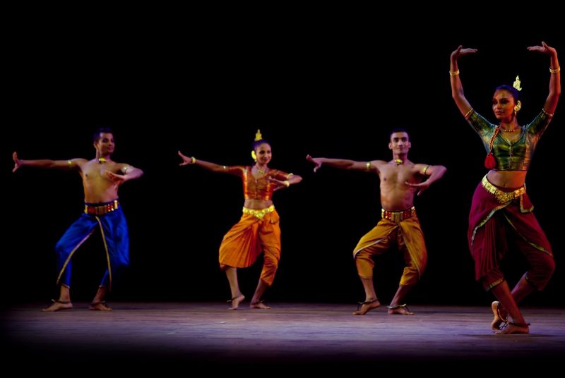 Performers with two dance ensembles perform onstage. A woman stands in front, wearing silky, flowing maroon pants and a silky green cropped top, gold jewelry on her wrists and neck. With her feet together, but her legs bent she reaches above her head with her hands. Behind her, three dancers move. One on the far left is a shirtless man with silky electric blue pants, the woman in the middle wears flowy orange pants with a matching cropped top, and a second shirtless man dances in silky yellow pants.