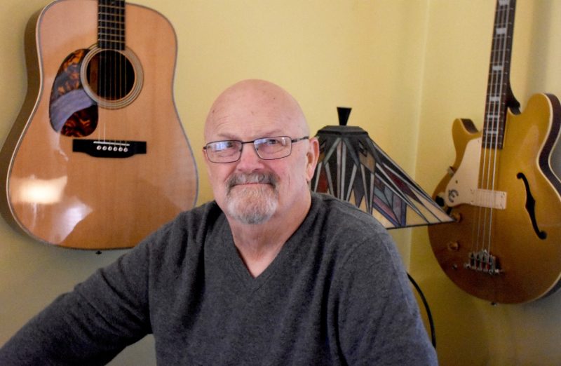 Robert Rogers, bald, with a short goatee, poses in his den, surrounded by guitars that are hung on the wall behind him.