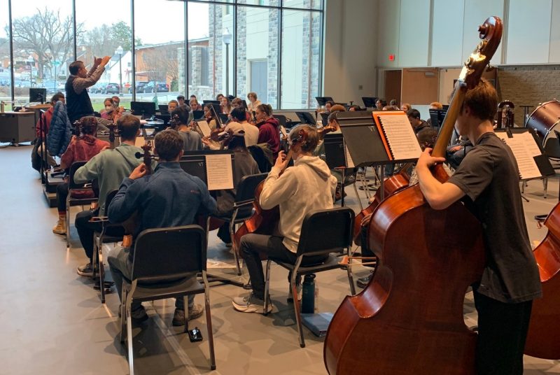 Students in the Virginia Tech Philharmonic orchestra practice in a rehearsal room with a large window, as they watch the conductor, standing in front of the group with his arms raised.