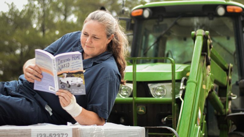 Veterinarian in blue overalls reading a book on a green tractor.