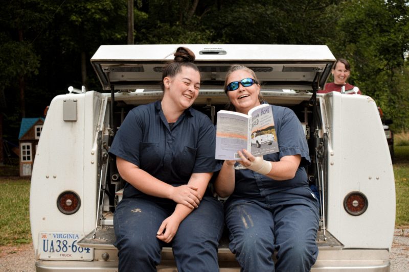Two veterinarians sitting on a white truck, in blue coveralls, reading a book.