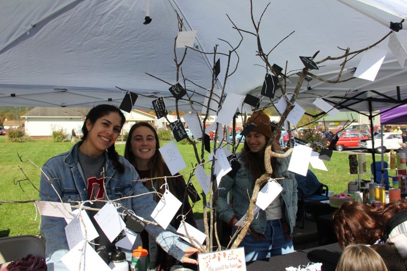 students outside under a tent, standing next to a story tree